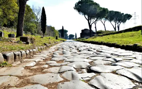 Ancient Roman cobblestone road lined with trees and ruins, leading towards the horizon.