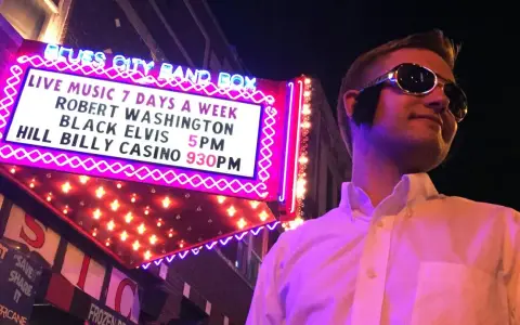 Mann im Elvis-Stil mit Sonnenbrille vor der "Blues City Band Box"-Neonreklame, die "Black Elvis" ankündigt.