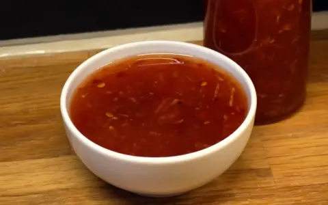 En skål med glansig, röd sweet chili sås med små bitar av chili och vitlök. En flaska med samma sås syns i bakgrunden.