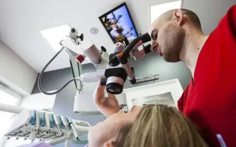Dentysta w czerwonej bluzie bada pacjentkę przez mikroskop stomatologiczny.