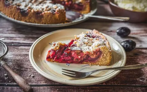 Ein saftiger Zwetschgenkuchen mit Dinkelmehl vom Blech, bestäubt mit Puderzucker. Ein Stück auf einem Teller mit einer Gabel.