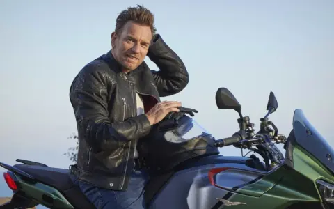 Ewan McGregor, con giacca di pelle, posa su una Moto Guzzi verde e grigia, pronto per un'avventura.