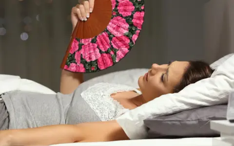 Frau liegt im Bett, nass geschwitzt, fächelt sich mit einem Blumenfächer Luft zu. Die Nacht ist heiß.