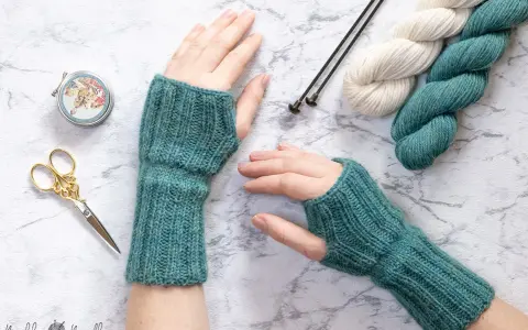 Manos con guantes tejidos a mano, listos para un nuevo patron. Lana, agujas y tijeras decorativas completan la escena.