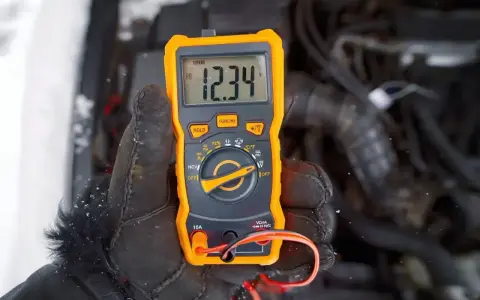 Ręka w rękawicy trzyma pomarańczowy multimetr, który pokazuje 12.34V. Auto nie odpala, akumulator sprawny, problem leży gdzie indziej.