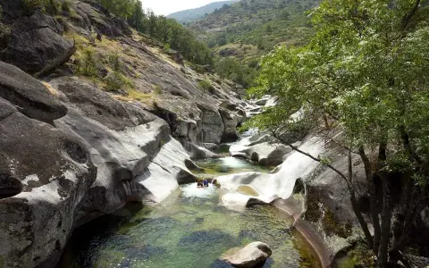 Un río de aguas cristalinas fluye entre rocas erosionadas, formando piscinas naturales. Dos personas disfrutan del baño en uno de los pilones, rodeados de vegetación y montañas.