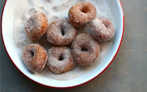 Små, nybakade donuts som rullats i socker och kanel ligger i en vit skål med röd kant.