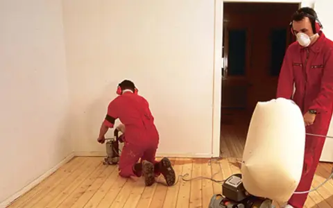 Zwei Männer in roten Overalls schleifen Dielen ab. Einer kniet und bedient eine Schleifmaschine, der andere steht und hält einen Staubfangsack.