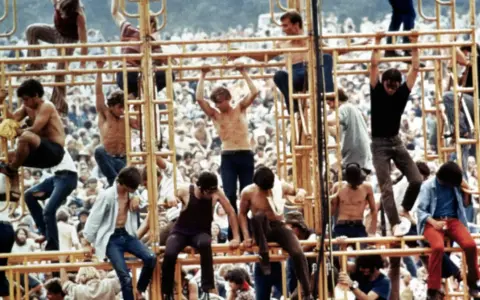 Tłum ludzi na festiwalu, niektórzy wspinają się na konstrukcję. To właśnie woodstock co to za wydarzenie?
