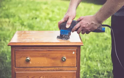 Un homme utilise une ponceuse pour le décapage mécanique d'une commode en bois, préparant le meuble pour une nouvelle finition.