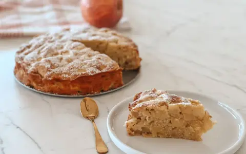 Une part de **crépiau aux pommes** saupoudrée de sucre glace, à côté d'un gâteau entier et d'une pomme. Une cuillère dorée est posée sur l'assiette.