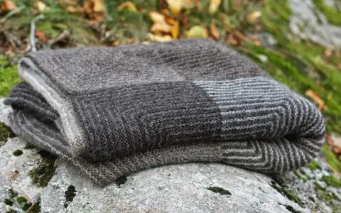 En hopvikt, handstickad filt i gråa nyanser ligger på en mossig sten. Perfekt för mysiga höstkvällar.