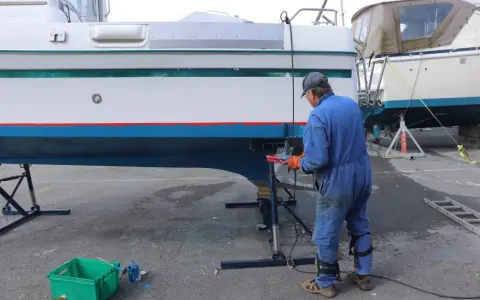 Un homme en combinaison bleue travaille sur la coque d'un bateau pour enlever l'antifouling. Des outils et des pots de peinture sont à proximité.