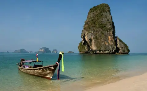 En traditionell longtailbåt flyter i turkost vatten vid en sandstrand i Thailand. Klippöar reser sig ur havet. Kanske en resa hit inkluderar vaccin Thailand pris.