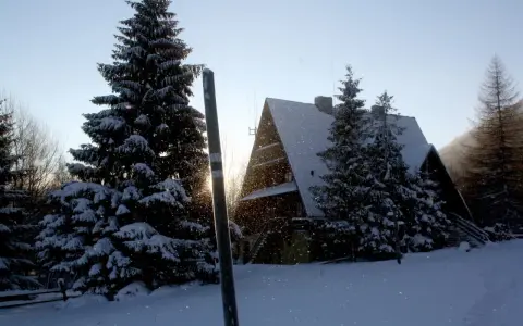 Zimowy krajobraz z bacówką pod Małą Rawką. Śnieg sypie, a słońce przebija się przez drzewa.