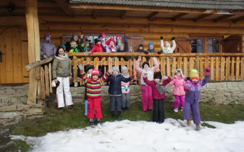 Wesoła gromadka dzieci pozdrawia z bacówki u harnasia. Wszyscy ubrani w zimowe stroje, radośnie machają rękami na tle drewnianego domu.