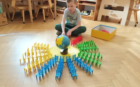 Chłopiec bawi się w przedszkolu Montessori, układając kolorowe krzesełka wokół globusa. To pokazuje, montessori co to za metoda nauczania.