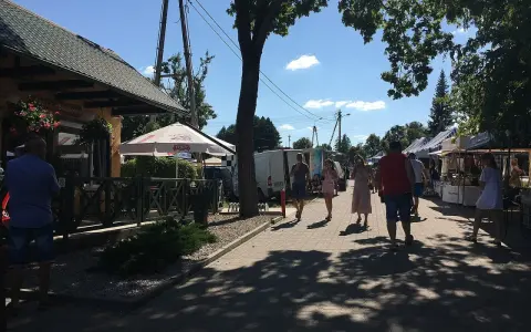 Ludzie spacerują po targu, podziwiając **tleń atrakcje**. Widać stoiska z towarami, restaurację i drzewa rzucające cień.
