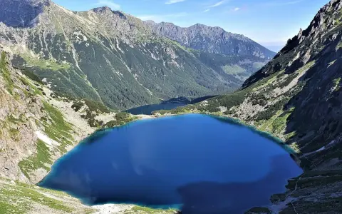 Widok na Dolinę 5 Stawów Wysokości. Głębokie, turkusowe jezioro otoczone stromymi, skalistymi zboczami gór porośniętych zielenią.