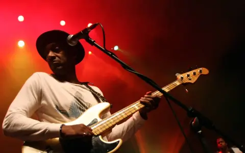 Marcus Miller w czarnym kapeluszu gra na białej gitarze basowej na tle czerwonych świateł scenicznych.