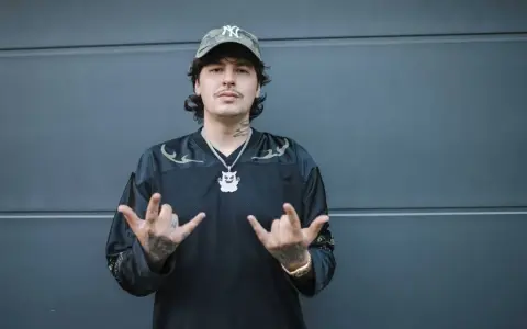 Młody mężczyzna w czapce z daszkiem i naszyjnikiem z wizerunkiem duszka, pozuje z uniesionymi dłońmi. Jego styl przypomina turystę z 2115 roku.