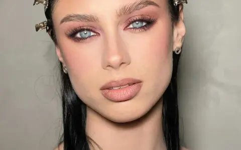 Ojos ahumados en tonos rosados y marrones, realzando unos ojos azules penetrantes. Labios nude y cabello oscuro completan el look.