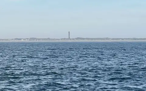 Der Leuchtturm Norderney ragt über die Dünenlandschaft am Horizont. Das weite Meer liegt davor.