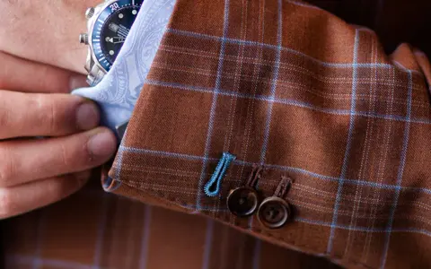 A man adjusts his watch on his wrist, revealing the detailed pick stitching on the cuff of his brown plaid suit jacket.