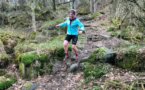 Kvinna springer i skogen, fokuserad på att springa marathon. Mossbeklädda stenar och träd skapar en utmanande terräng.