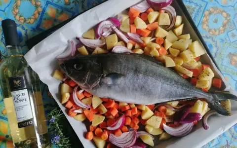 Ricciola al forno pronta per essere infornata con patate, carote e cipolle rosse. Accanto, una bottiglia di vino bianco.