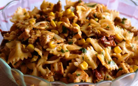 Pyszna sałatka z makaronem farfalle, kurczakiem, kukurydzą i suszonymi pomidorami, posypana natką pietruszki.