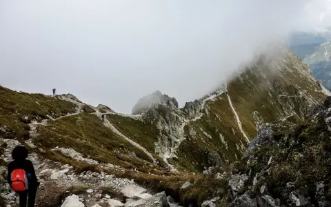 Mglista pogoda na Szpiglasowym Wierchu. Turysta z czerwonym plecakiem na szlaku wśród kamieni i traw.