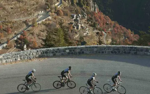 Ciclisti affrontano la salita del Col de la Couillole, con un villaggio arroccato sulla montagna autunnale.