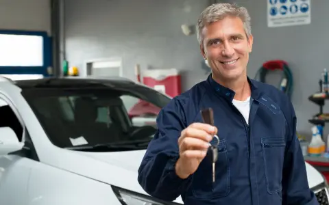 Uśmiechnięty mechanik w granatowym kombinezonie podaje kluczyki do białego samochodu. W tle warsztat samochodowy, gdzie wszystko jest gotowe, by sprawdzić, co to homologacja.