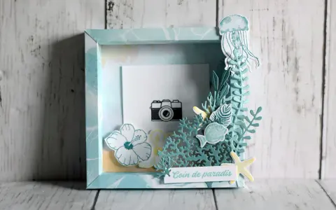 Un cadre photo en bois décoré sur le thème marin avec une méduse, des poissons, des fleurs et une petite caméra.