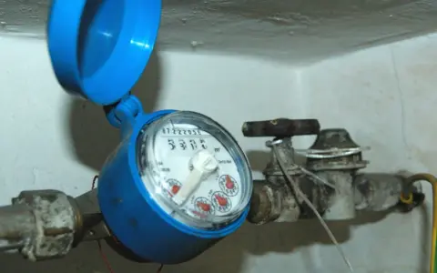 Contatore acqua blu con coperchio aperto, il quadrante è illeggibile per condensa. Tubi e rubinetto arrugginiti.
