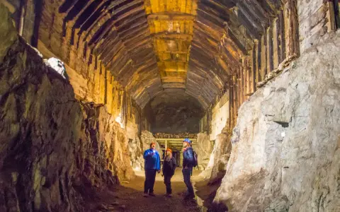 Zwiedzanie Kompleksu Riese: trzej turyści w kaskach oświetlają latarkami mroczny, drewniany tunel.