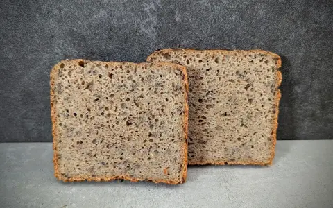 Zwei Scheiben rustikales **Sauerteig Kastenbrot** mit Körnern und einer luftigen Krume vor dunklem Hintergrund.