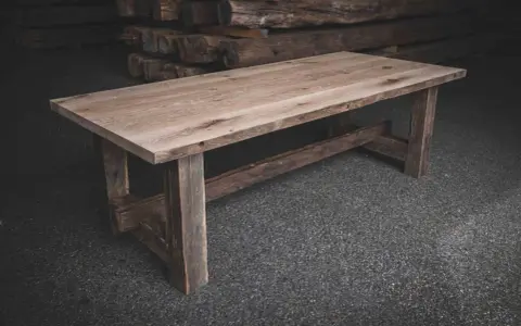 Ett rustikt bord av oljad ek med grova ben och en bred bordsskiva. Bakom syns staplade trästockar.