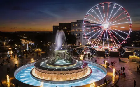 Una piazza illuminata a festa a Malta a Natale, con una fontana scintillante e una ruota panoramica che domina lo skyline al tramonto.