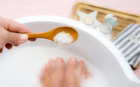 A hand adds epsom salt and baking soda to a foot soak, with feet submerged in the milky water.