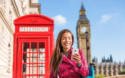 Uśmiechnięta kobieta z telefonem przed czerwoną budką telefoniczną i Big Benem. Pyta, czy w UK jest roaming, bo chce zadzwonić do domu.