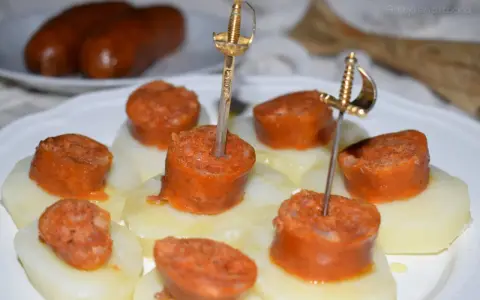 Chorizo a la sidra en rodajas, servido sobre patatas y adornado con espadas doradas.