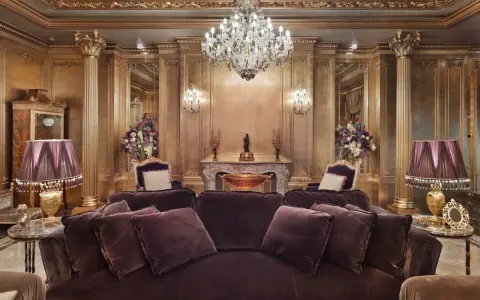 Salón lujoso con sofás de terciopelo morado, lámparas doradas y un candelabro de cristal. El techo y las paredes doradas evocan el estilo barroco español.