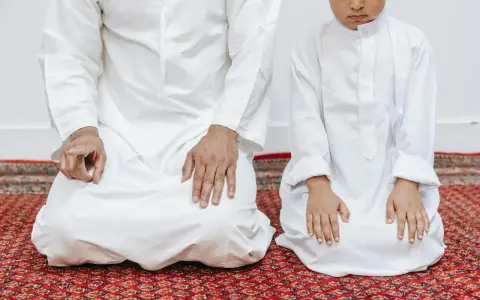 Ojciec i syn w białych strojach klęczą na dywanie, wykonując codzienną modlitwę poranną.