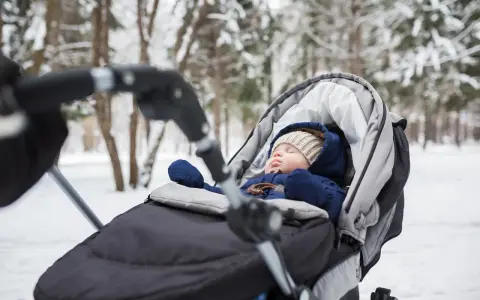 Śpiące dziecko w wózku zimą. Ciepły kombinezon, czapka i rękawiczki to podstawa, co włożyć do wózka dla noworodka zimą.