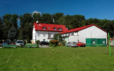 Budynek z czerwonym dachem, przypominający dawny korsarz darłówko, otoczony zielenią i samochodami.
