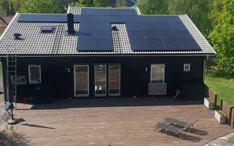 Ett mörkt hus med en liten solcellsanläggning på taket. En stor altan med solstolar leder ut mot en grönskande trädgård.