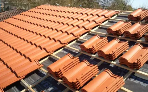 Vue d'un toit en cours de pose, révélant des rangées de tuiles mécaniques en terre cuite orange vif, empilées sur des lattes de bois.