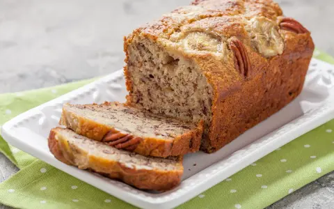Pyszny chlebek bananowy z 2 bananów, z orzechami pekan i plasterkami banana na wierzchu, na białej paterze.